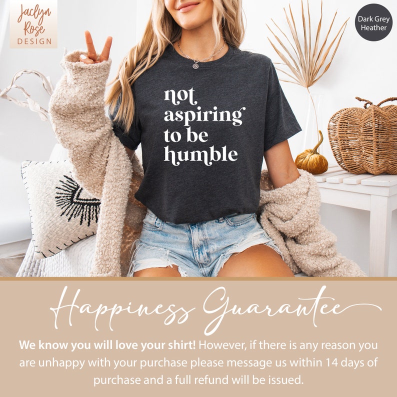 May include: A dark grey heather t-shirt with the text "not aspiring to be humble" printed in white. The shirt is being worn by a person with long blonde hair. The text "Happiness Guarantee" is printed in a cursive font on a light brown background.