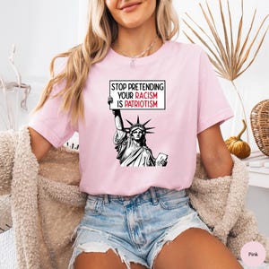 Peut inclure: Un t-shirt rose avec un graphique de la Statue de la Libert&eacute; tenant une pancarte qui dit "Arr&ecirc;tez de pr&eacute;tendre que votre racisme est du patriotisme".