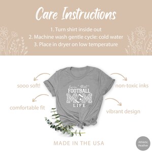May include: Gray t-shirt with the text "livin' that FOOTBALL MOM LIFE" in white. The shirt is made in the USA and features a comfortable fit, non-toxic inks, and a vibrant design.