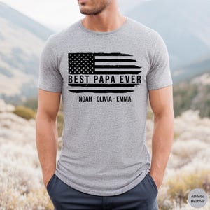 May include: A gray t-shirt with a distressed American flag design and the text "Best Papa Ever" with the names "Noah - Olivia - Emma" underneath.