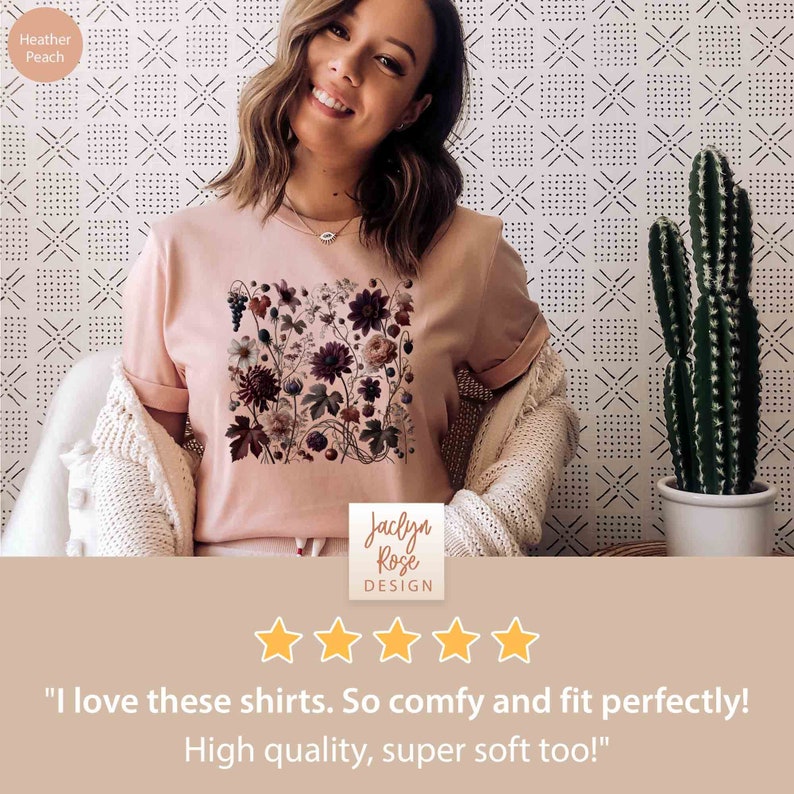 Op de afbeelding: Een vrouw draagt een heideperzikkleurig t-shirt met een bloemenprint. Het shirt heeft een ronde hals en korte mouwen. De bloemenprint bevat een verscheidenheid aan bloemen, bladeren en bessen in tinten roze, rood en groen. De tekst "Jaclyn Rose DESIGN" is gedrukt op de onderkant van het shirt. De vrouw glimlacht en kijkt naar de camera. De achtergrond is een witte muur met een zwart-wit patroon.