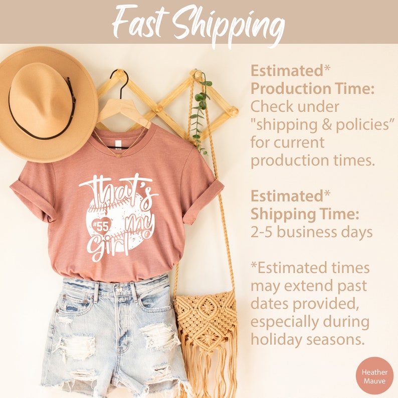 May include: A heather mauve t-shirt with a white baseball graphic and the text "That's my girl" with the number 55 inside the baseball. The shirt is hanging on a wooden rack with a straw hat and a woven bag.