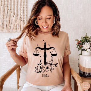May include: A light pink t-shirt with a black Libra zodiac symbol, a scale with stars, and floral accents. The text "LIBRA" is printed below the symbol.