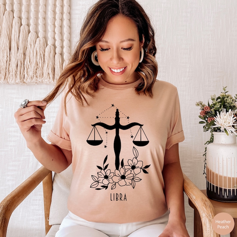 May include: A light pink t-shirt with a black Libra zodiac symbol, a scale with stars, and floral accents. The text "LIBRA" is printed below the symbol.