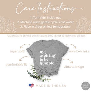 May include: A gray t-shirt with the text "not aspiring to be humble" printed in white. The shirt is made in the USA and is super soft, comfortable, and features a vibrant design. The graphics are printed using a direct-to-garment process with non-toxic inks.