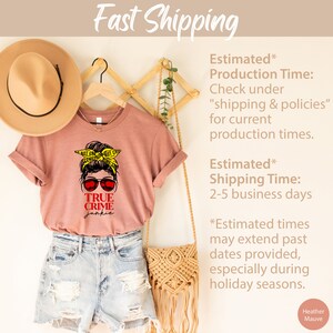 May include: A heather mauve t-shirt with a graphic of a woman with a messy bun wearing sunglasses and a yellow crime scene tape headband. The text "TRUE CRIME junkie" is printed in red below the graphic. The shirt is hanging on a wooden hanger with a straw hat and a woven bag.