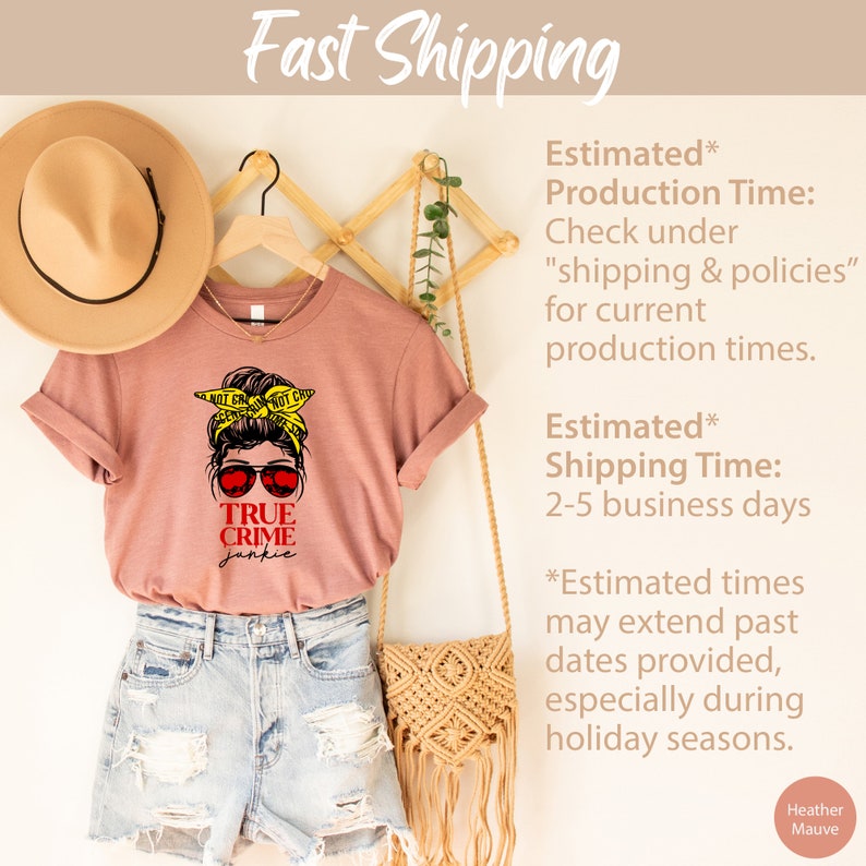 May include: A heather mauve t-shirt with a graphic of a woman with a messy bun wearing sunglasses and a yellow crime scene tape headband. The text "TRUE CRIME junkie" is printed in red below the graphic. The shirt is hanging on a wooden hanger with a straw hat and a woven bag.