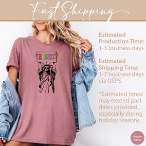 May include: A heather mauve t-shirt featuring a black and white illustration of the Statue of Liberty holding a sign that says "RESIST" in rainbow colors.
