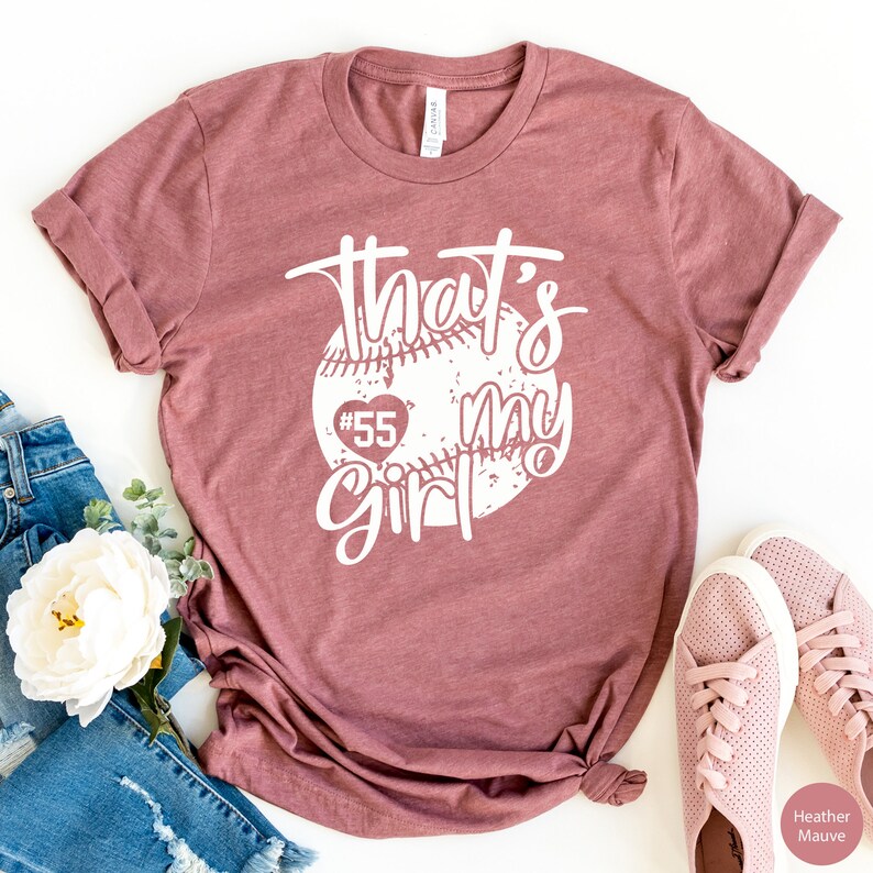 May include: A heather mauve t-shirt with a white graphic of a baseball with a heart and the text "That's my girl #55".