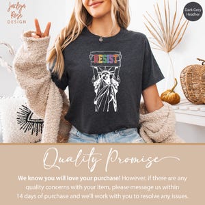 May include: A dark grey heather t-shirt featuring a white outline of the Statue of Liberty holding a sign that says "RESIST" in rainbow colors.
