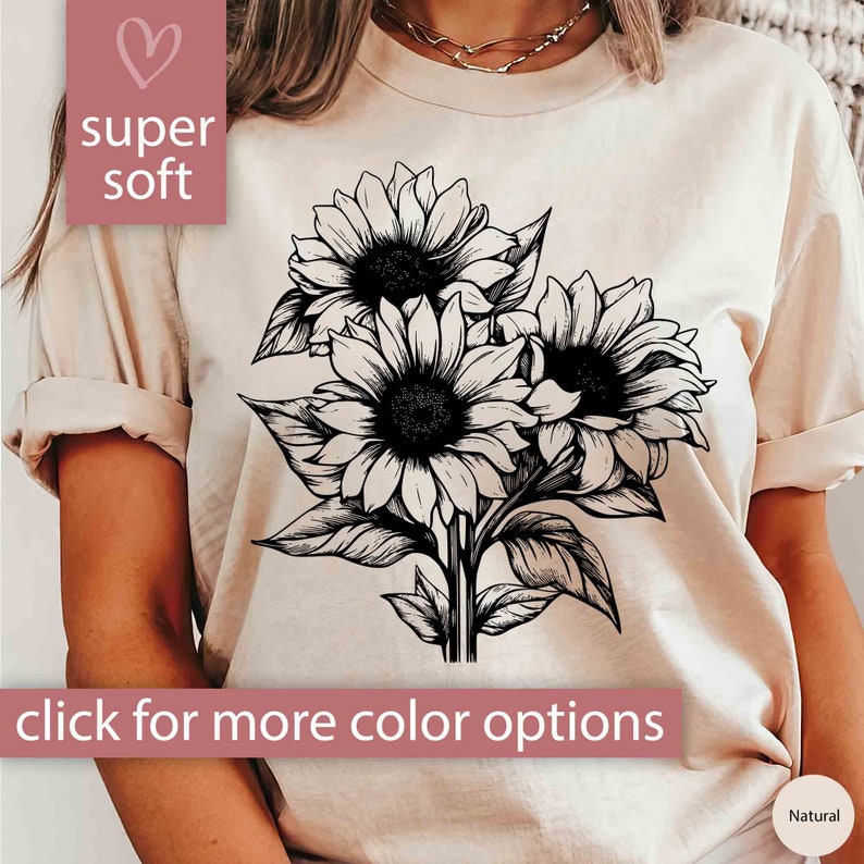May include: A beige t-shirt featuring a black and white illustration of three sunflowers. The words "super soft" and "click for more color options" are displayed on the shirt. The shirt is labeled "Natural".
