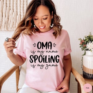 May include: Pink t-shirt with black text that reads "OMA is my name SPOILING is my game".