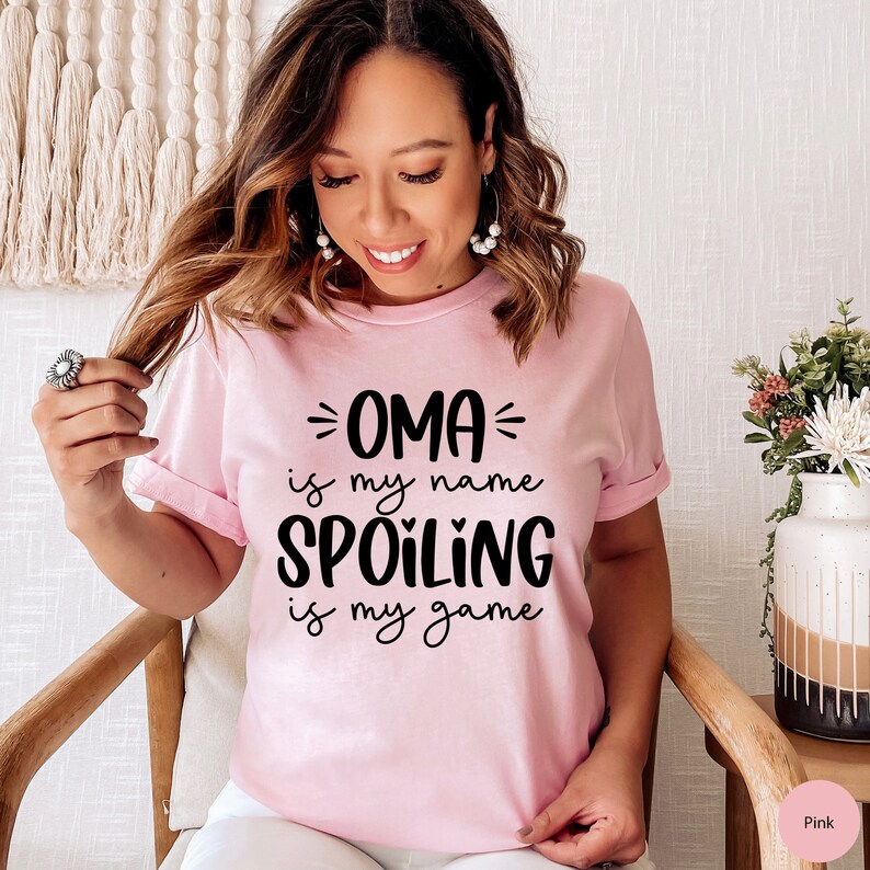 May include: Pink t-shirt with black text that reads "OMA is my name SPOILING is my game".