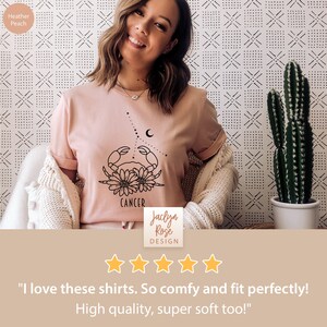 May include: A pink t-shirt with a black outline of a crab and flowers, with the word "Cancer" printed below. The shirt is worn by a person with a smiling face. The text "Jaclyn Rose DESIGN" is printed below the image. Five gold stars are displayed below the text. The text "I love these shirts. So comfy and fit perfectly! High quality, super soft too!" is printed below the stars.