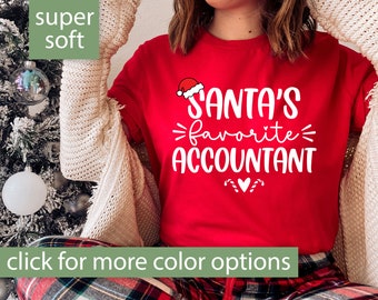 May include: Red t-shirt with white text that says "Santa's favorite accountant" with a small Santa hat graphic above the text. The shirt is being worn by a person with a white cardigan and red and green plaid pants.