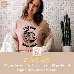 Puede incluir: Una camiseta rosa con un dise&ntilde;o gr&aacute;fico negro de una luna creciente, hongos y flores. El texto "Stay Wild Moon Child" est&aacute; impreso en la camiseta. La camiseta est&aacute; siendo modelada por una persona. El texto "Jaclyn Rose Design" est&aacute; impreso en una etiqueta blanca unida a la camiseta.