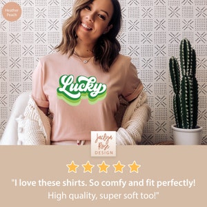 May include: A woman wearing a pink t-shirt with a green "Lucky" graphic. The shirt is a soft, comfortable material. The t-shirt is on a white background with a cactus in the background. The text "Jaclyn Rose DESIGN" is on the bottom of the image. The image has a five-star rating and the text "I love these shirts. So comfy and fit perfectly! High quality, super soft too!"