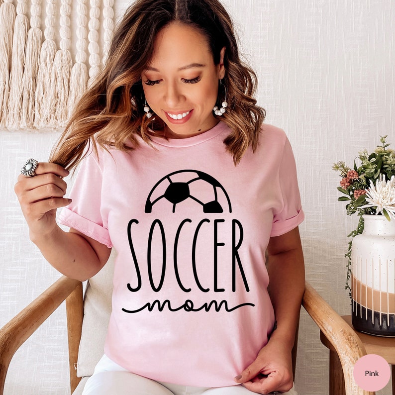 May include: Pink t-shirt with a black soccer ball graphic and the text "soccer mom" in black.