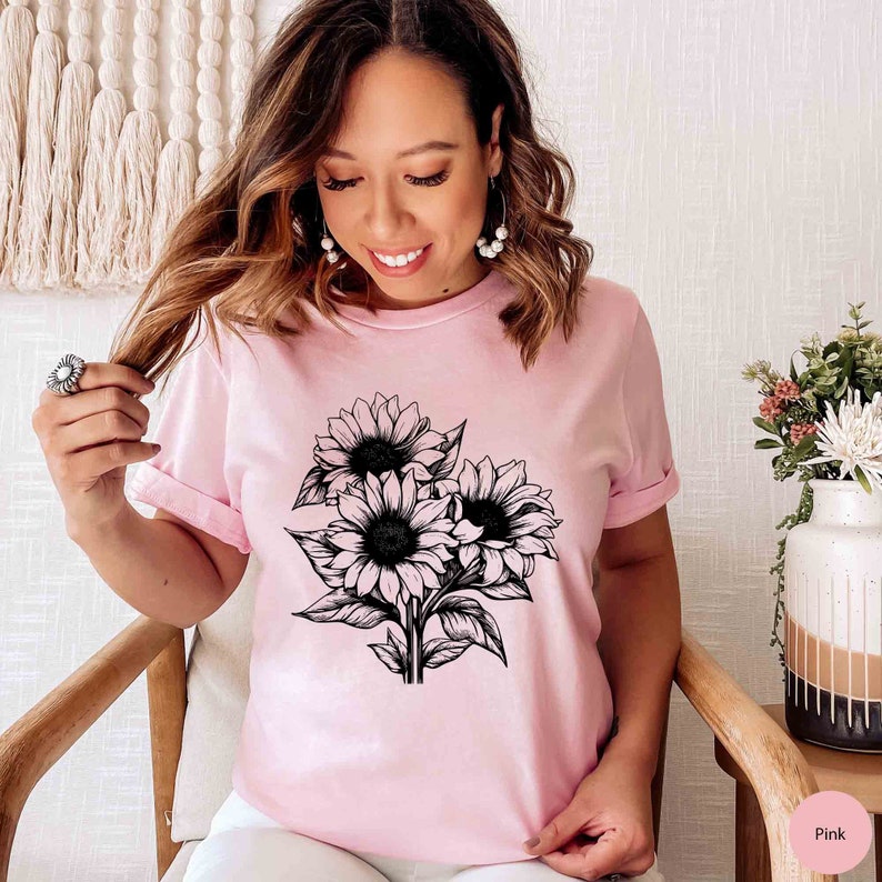 May include: A light pink t-shirt featuring a black and white sunflower bouquet design. The graphic shows three sunflowers with detailed petals and leaves. The shirt has short sleeves and a classic crew neck.