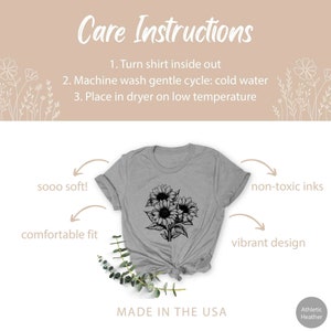 May include: Athletic Heather gray t-shirt with a black sunflower design. The shirt has the text "Care Instructions" and "MADE IN THE USA". Additional text includes "sooo soft!", "comfortable fit", "non-toxic inks", and "vibrant design".