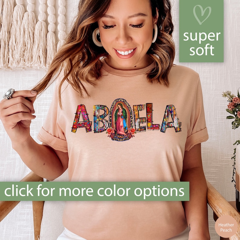 May include: A light peach colored t-shirt with the word "ABUELA" spelled out in colorful letters. The letters are decorated with a variety of patterns and designs, including flowers, religious imagery, and geometric shapes. The word "ABUELA" is centered on the shirt. The shirt is made of a soft, comfortable fabric.