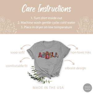 May include: A gray t-shirt with the word "ABUELA" spelled out in colorful mosaic letters. The letters are decorated with images of flowers, birds, and a religious figure. The shirt is made in the USA and features a comfortable fit, soft fabric, and non-toxic inks.