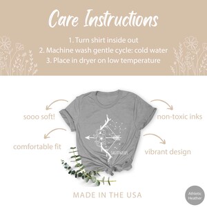 May include: Grey t-shirt featuring a white design of an arrow and flowers with the word "SAGITTARIUS" underneath. The shirt is made in the USA and is described as "sooo soft!" and "comfortable fit". The ink is non-toxic and the design is vibrant.