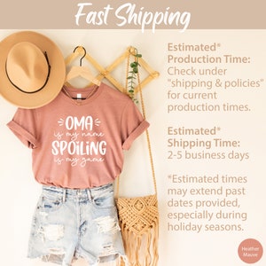 May include: A heather mauve t-shirt with white text that reads "OMA is my name SPOILING is my game". The shirt is hanging on a wooden hanger with a straw hat and a woven bag.