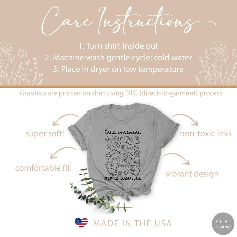 Op de afbeelding: Athletic heather grijs t-shirt met de tekst "less meanies more weenies" en een zwarte hondenillustratie. Het shirt is gemaakt in de VS en bevat onderhoudsinstructies. Het wordt beschreven als superzacht, comfortabel en met een levendig ontwerp.