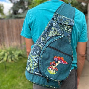 May include: A teal blue sling backpack with a psychedelic pattern and an embroidered mushroom design. The backpack has a zippered pocket on the front and a smaller pocket on the side.