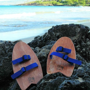 May include: Two wooden surfboard-shaped sandals with blue straps. The sandals are on a rocky beach with the ocean in the background. The sandals have the text "Made with Aloha" printed on them.