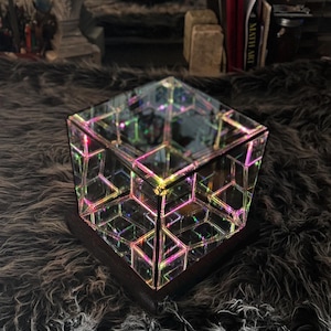 May include: A geometric, mirrored cube with internal, colorful light patterns. The cube sits on a dark wooden base, reflecting a kaleidoscope of colors. The background is a dark, textured surface.