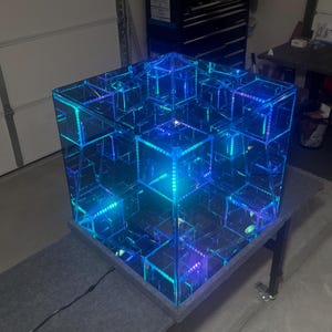 May include: A large, illuminated cube with a mirrored interior, creating an infinity effect. The cube is lit with blue and teal LED lights, giving it a futuristic appearance. The cube sits on a gray surface.