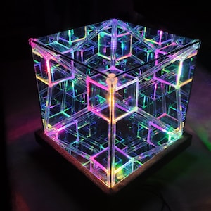 Può includere: Una lampada a cubo infinito quadrata con una superficie a specchio riflettente e luci LED interne che creano un motivo geometrico ripetitivo di linee luminose color rosa neon, blu e verde all'interno di una cornice trasparente su una base scura.