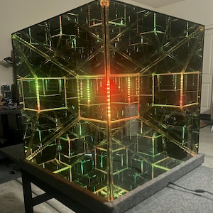 May include: A large, mirrored cube with internal reflections and colorful LED lighting. The cube's interior features a complex geometric pattern, with red, green, and yellow lights creating a mesmerizing visual effect. The cube sits on a dark surface.