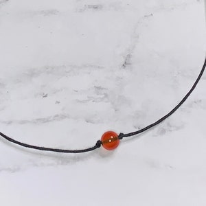 May include: A simple necklace with a single, round, orange gemstone bead on a black cord.