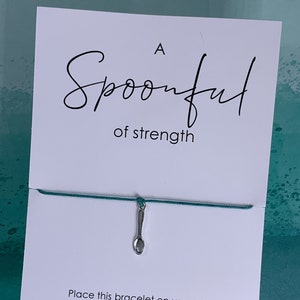 May include: A white card with the text "A Spoonful of strength" and a green string bracelet with a silver spoon charm. The card instructs the user to place the bracelet on their wrist, tie a knot, and make a wish.