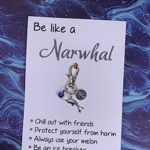 May include: A white card with the text "Be like a Narwhal" and a list of bullet points: "Chill out with friends", "Protect yourself from harm", "Always use your melon", "Be an ice breaker", "Dive deep into life", "Come up for air". A silver narwhal keychain with a blue crystal charm is attached to the card.
