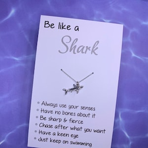 May include: White card with the text "Be like a Shark" and a silver shark necklace. The card lists the following qualities of a shark: Always use your senses, Have no bones about it, Be sharp & fierce, Chase after what you want, Have a keen eye, Just keep on swimming.