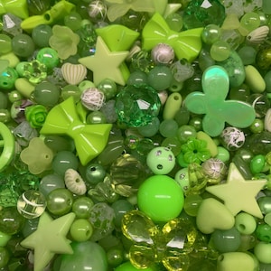 May include: A collection of green beads and embellishments in various shades, including lime, olive, and emerald. The assortment features round beads, star shapes, flower shapes, and bow shapes, perfect for jewelry making or craft projects.
