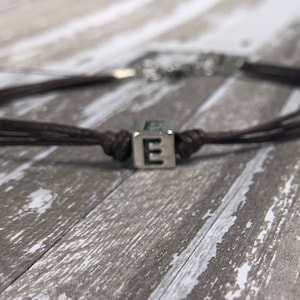 May include: A brown leather cord bracelet with a silver charm featuring the letter "E".
