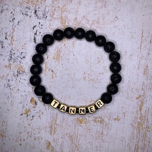 May include: Black lava rock beaded bracelet with gold letter beads spelling the name "TANNER".