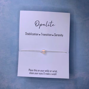 May include: White string bracelet with a small, round opalite bead. The card reads "Opalite", "Stabilization. Transition. Serenity", and "Place this on your ankle or wrist, close your eyes & make a wish!"