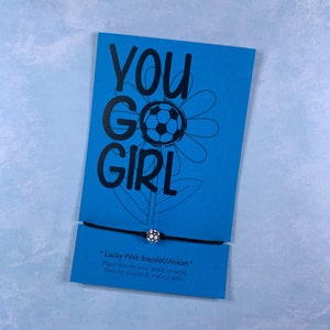 May include: A blue card with the text "YOU GO GIRL" in black lettering. A black and white soccer ball graphic is in the center of the card. A black string bracelet with a white and black soccer ball charm is attached to the card. The card reads "Lucky Wish Bracelet/Anklet* Place this on your ankle or wrist, then tie a knot & make a wish!"