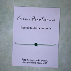 May include: Green aventurine bracelet with a green bead on a white card with the text "Green Aventurine Opportunity. Luck. Prosperity Place this on your ankle or wrist, close your eyes & make a wish!"