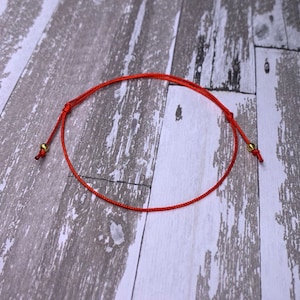 May include: A red string bracelet with gold beads at each end. The bracelet is tied in a knot and is lying on a wooden surface.