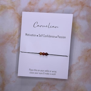 May include: A white card with the word "Carnelian" in script, and the words "Motivation, Self Confidence, Passion". A black string with three orange-brown beads is attached to the card. The text "Place this on your ankle or wrist, close your eyes & make a wish!" is also on the card.