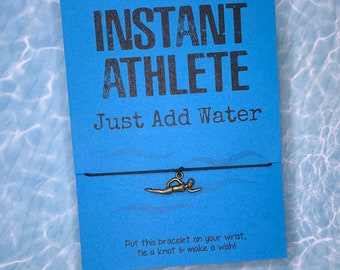 May include: A blue card with the text "INSTANT ATHLETE Just Add Water" and a black string bracelet with a gold swimmer charm. The card also says "Put this bracelet on your wrist, tie a knot & make a wish!"
