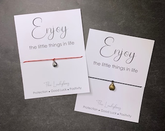 May include: Two cards with the text "Enjoy the little things in life" and a red and black string bracelet with a ladybug charm. The charm is silver on the red bracelet and gold on the black bracelet. The cards also have the text "The Ladybug Protection. Good Luck. Positivity."