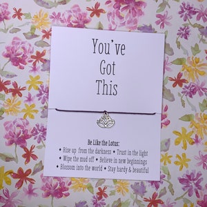 May include: A white card with the text "You've Got This" and a silver lotus charm attached to a purple string. The card also includes the text "Be Like the Lotus:" and a list of inspirational quotes about the lotus flower.